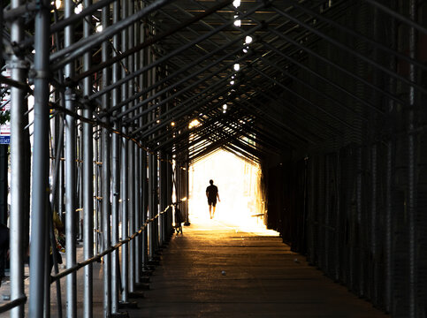 Man walking down the city sidewalk under construction scaffolding with light glowing in the background © deberarr