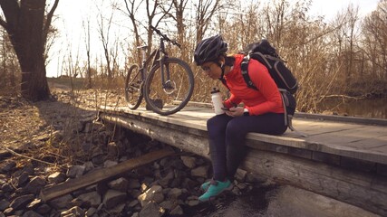 Happy woman sits wooden bridge over river, listening music in headphones with smartphone hands and drinks water white bottle. cyclist listens song in nature. athlete near bike hand on plastic drink