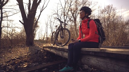 Happy young woman sitting wooden bridge over river, listening music in headphones with smartphone hands and dancing. Positive cyclist listens song in nature. Athlete happy near bike ears earphones