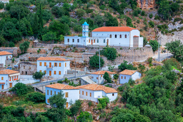 Fototapeta na wymiar View of Albanian village - traditional white houses with orange roofs and wooden shutters on windows - on the mountain hill, small church and road.