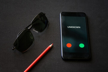 A smartphone with an unknown call lies on a black surface next to a red pencil and sunglasses. The...