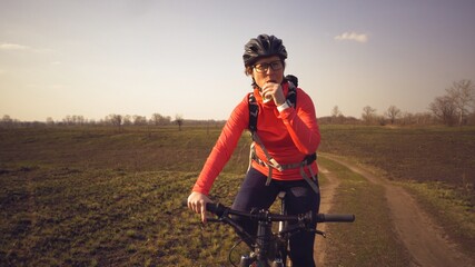 Athletic Caucasian woman eats protein bar ride on mountain bike on nature. Young sporty woman athlete in helmet resting while biting nutritional bar. Fitness woman eating energy snack outdoor