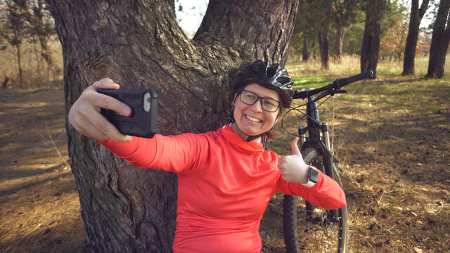 Young caucasian woman athlete tourist cyclist uses hand smart phone photo of herself selfie sitting near tree in coniferous forest outside the city. Sportswoman taking selfie with her mountain bike