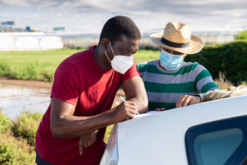 Farmer and worker in medical masks signing papers standing near car on farm field. Pandemic prevention and social distancing concept