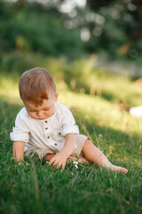 Happy adorable baby boy sitting on the grass in the park on summer day. Child in trendy and cute clothes