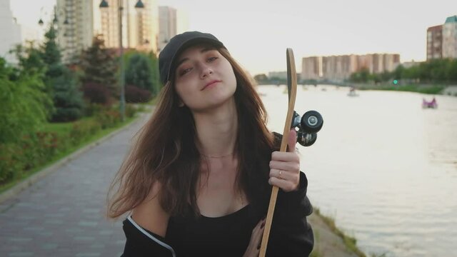 Beautiful and fashionable young woman posing with a skateboard on river embankment