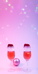 Christmas and new year holiday greeting card concept with two wine glasses with rose wine with Santa hat bauble on pink background in neon lights with multicolored snowflake bokeh. Copy space.