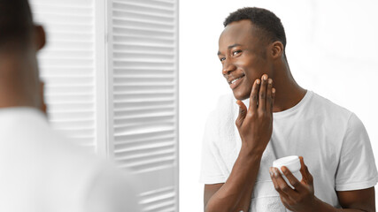 Fototapeta na wymiar Black Man Using Moisturizing Facial Cream Standing In Bathroom Indoor