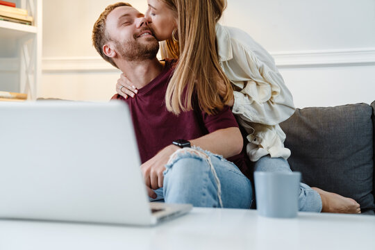 Image of ginger couple kissing and using laptop while sitting on sofa