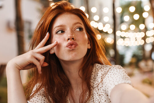 Image of cheerful ginger girl gesturing peace sign while taking selfie