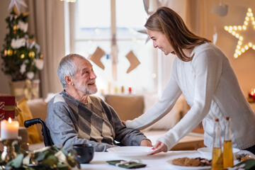 Young woman with senior grandfather in wheelchair indoors at home at Christmas.