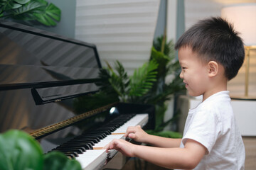 Cute happy smiling little Asian kid boy playing piano in living room at home, Preschool child...