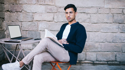 Thoughtful male freelancer in glasses working with documents and computer remotely in street cafe
