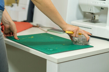 Seamstress's desk - female hands hold cloth cutting tools - knife and pins, other sewing accessories are on the table, a piece of orange cloth and white electric sewing machine in the background in bl