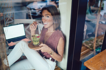 Cheerful smiling woman in casual wear enjoying work break holding glass with healthy tasty green...