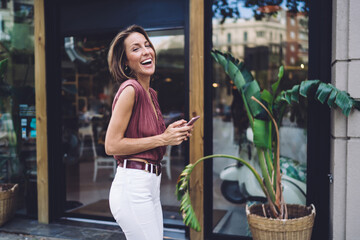 Half length portrait of cheerful caucasian woman n trendy wear walking on street with smartphone enjoying leisure, smiling pretty female blogger influencer satisfied with successful online job