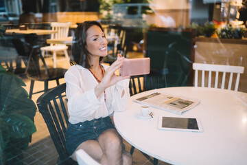 Cheerful adult woman taking selfie on smartphone in cafe