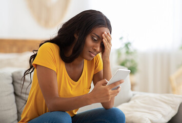 Unhappy African American lady looking at her smartphone with disgust indoors, copy space