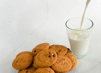 Milk is poured into a glass from above in a thin stream, next to oatmeal cookies with chocolate chips