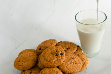 Milk is poured into a glass from above in a thin stream, next to oatmeal cookies with chocolate chips. White marble background. Selective focus. Copy space. Banner