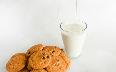 Milk is poured into a glass from above in a thin stream, next to oatmeal cookies with chocolate chips. White marble background. Selective focus. Copy space. Banner