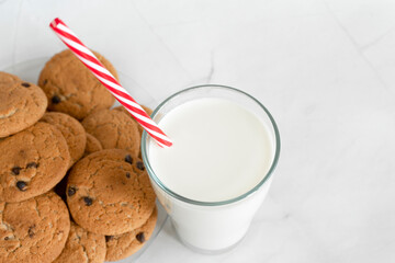 Milk in a glass with a straw, next to oatmeal cookies with chocolate chips close-up. Christmas drinks. White marble background. View from above. Selective focus. Copy space. Banner