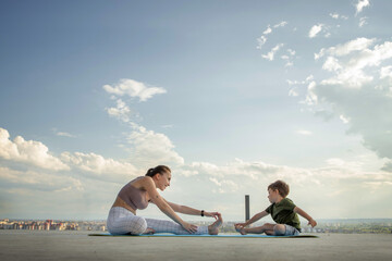 Fototapeta premium Mother and son doing exercise on the balcony in the background of a city during sunrise or sunset, concept of a healthy lifestyle