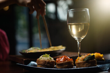 Close up shot of female hand enjoying Japanese cuisine various side dishes by chopstick, inari sushi set and white wine in restaurant with sun rays