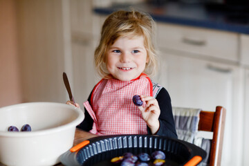 Cute little toddler girl baking plum pie at home. Happy smiling child helping and preparing plums for cake in domestic kitchen. Healthy homemade food.