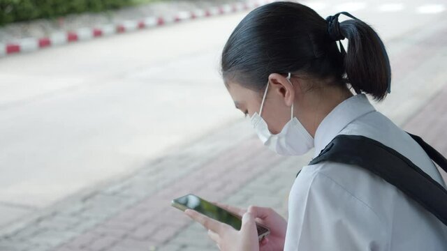 An Asian female high school student in white uniform wearing the mask use smartphone and sit , wait for car to go back home after school during the Coronavirus 2019 (Covid-19) epidemic.