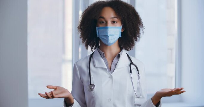 Female African American doctor nurse in medical mask standing in hospital clinic, speaking with patient on camera online video conference chat, gesticulate with hands, virus concept, webcam view