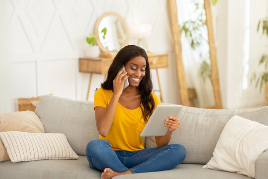 Black lady using tablet computer for online work, education or entertainment while talking on smartphone at home