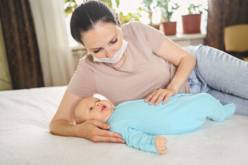 Obraz na płótnie Canvas Beautiful mother in protective face mask holding her little cute newborn son protecting him from viruses and infections. Coronavirus covid-19 prevention. Home quarantine. Happy family at home.