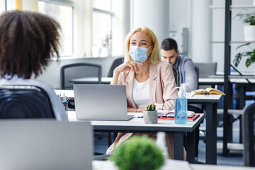 Communication of colleagues in office or client consultation and social distance. Millennial woman in protective mask talking with african american lady at workplace