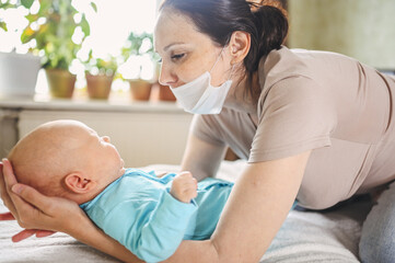 Obraz premium Beautiful mother in protective face mask holding her little cute newborn son protecting him from viruses and infections. Coronavirus covid-19 prevention. Home quarantine. Happy family at home.