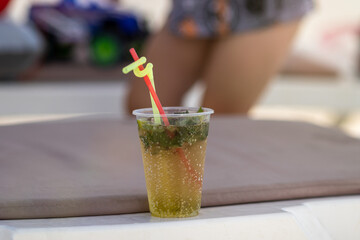 Mojito in a plastic cup with a yellow and red straw on the beach on a bright sunny day. Close-up with blurred background.