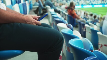 A man sits on the stadium stand and surfing social media on his smart phone during a football or soccer match. Mass event during coronavirus quarantine. Close-up of hands. High quality 4k footage - Powered by Adobe