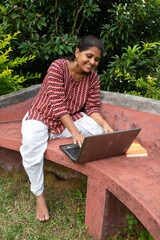 Beautiful happy adult woman using a Laptop on a red bench at a park with an orange book beside her.