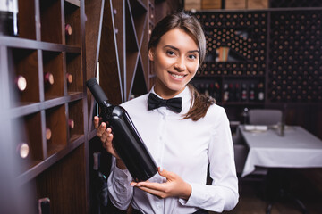 Selective focus of sommelier holding bottle of wine and looking at camera near racks - obrazy, fototapety, plakaty