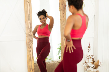 Fototapeta na wymiar Joyful Black Lady After Weight Loss Posing Near Mirror Indoors