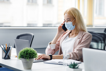 New normal and return to workplace after quarantine.Young attractive woman in a protective mask talks on smartphone and works in laptop, at table with antiseptic