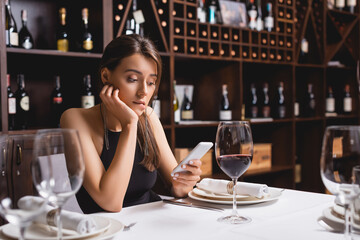Selective focus of young woman using smartphone while sitting near glass of wine in restaurant