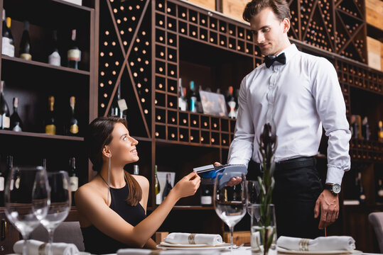 Selective focus of young woman holding credit card near waiter in formal wear with payment terminal in restaurant