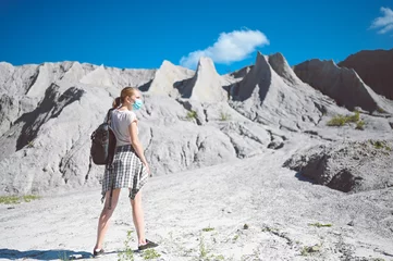 Fotobehang Young blonde woman walking against the white mountains wearing face protective mask for Covid 19 prevention. Young student traveler tourist with backpack. Corona virus and domestic tourism concept © Алина Троева