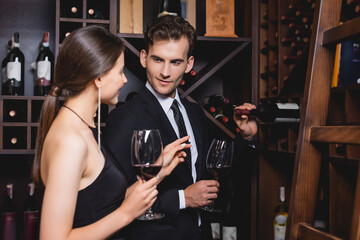 Selective focus of man in formal wear taking bottle from rack near elegant girlfriend with glass of wine