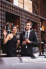 Selective focus of elegant woman waving hand while holding glass of wine near boyfriend in restaurant