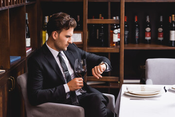 Selective focus of man in suit looking at wristwatch and holding glass of wine in restaurant