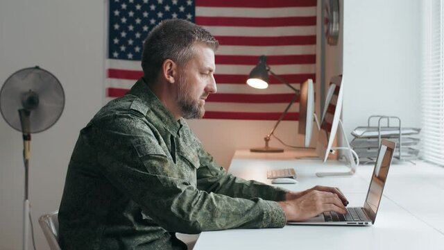 Slowmo side view shot of serious middle-aged U.S. army officer in military uniform sitting at desk in office and working on laptop