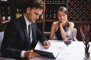 Selective focus of man in suit looking at menu near glasses of wine during dating with girlfriend in restaurant