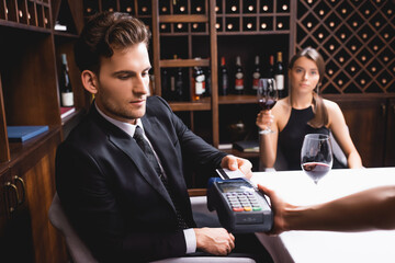 Selective focus of man with credit card paying to waitress with payment terminal during dating with girlfriend in restaurant